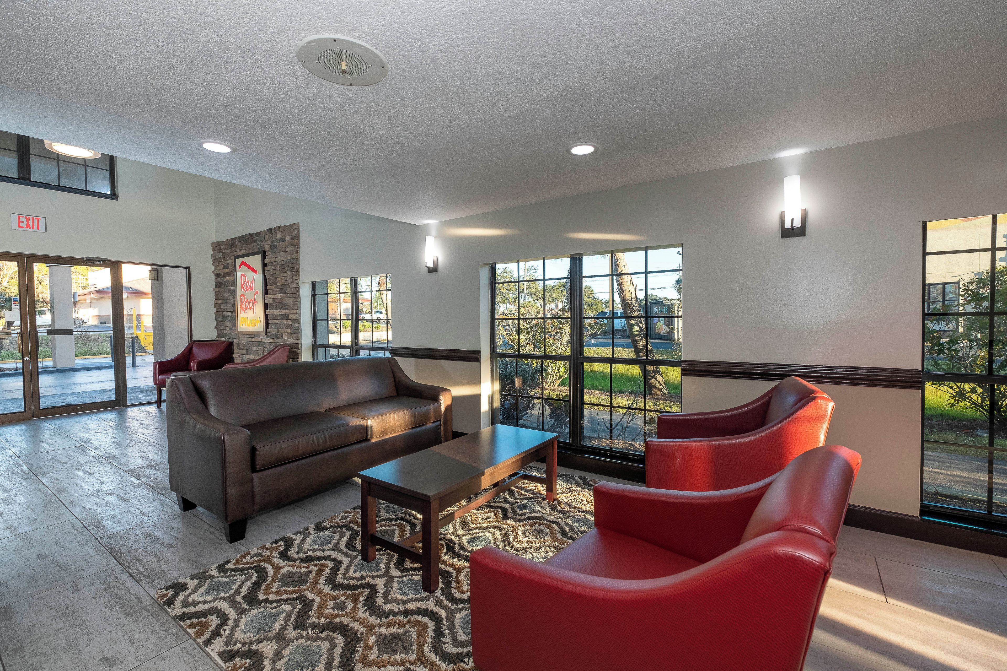 A lobby with a brown leather sofa, a wooden coffee table, and red chairs. There are large windows with a view of greenery and a building outside.