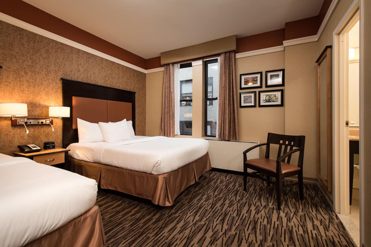 A hotel room with two beds, a sitting area with a chair, a desk with a lamp, and a window with curtains.