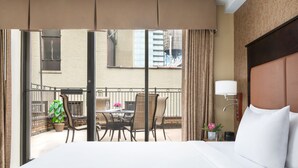 A hotel room with a bed, a nightstand, a lamp, and a view of an outdoor balcony with a table and chairs. - The Hotel @ Fifth Avenue (New York)