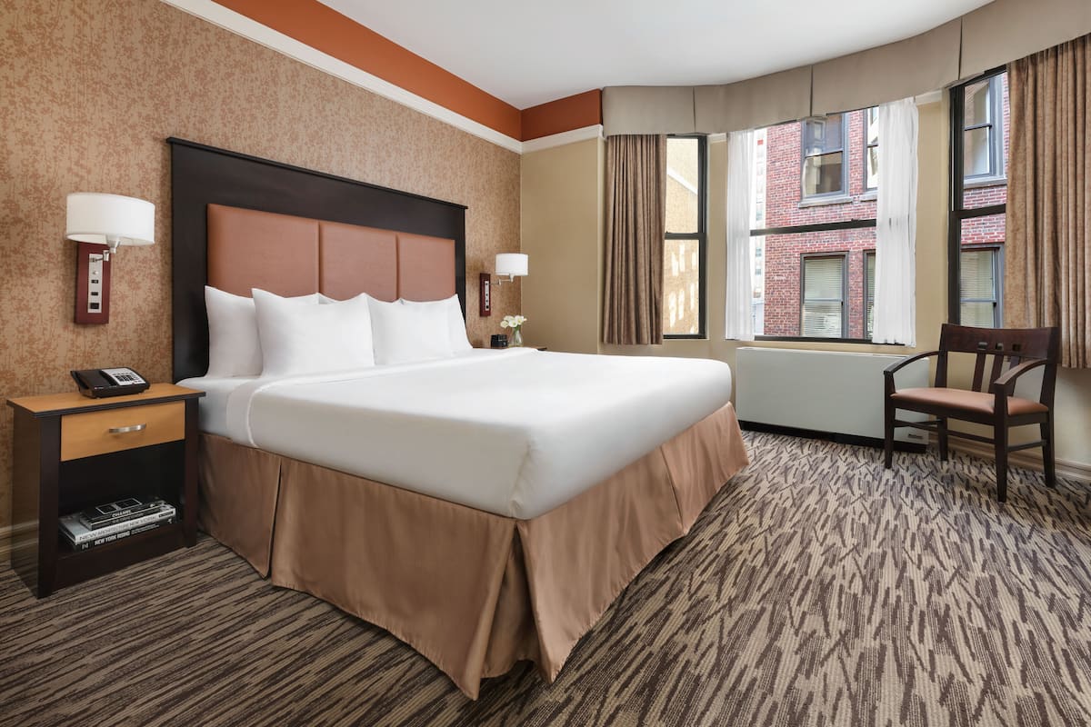 A hotel room with a large bed, a nightstand with a phone, a chair, and a window with curtains.