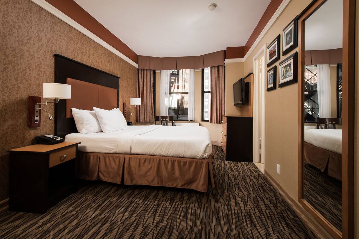 A hotel room with a large bed, a nightstand with a phone, a desk lamp, a mirror, and a view of the outside.
