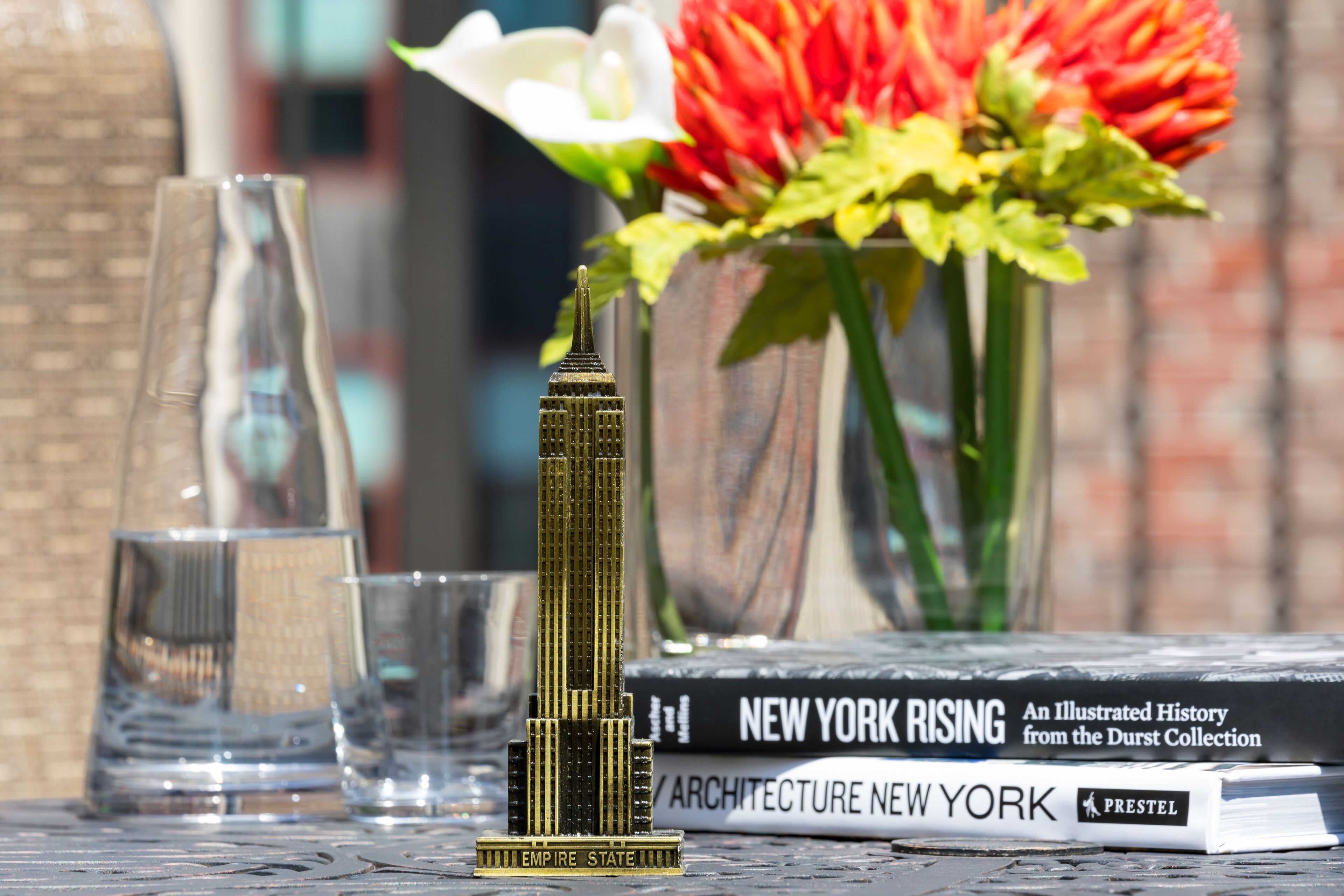 A model of the Empire State Building, a glass vase with flowers, and a book titled 'New York Rising' on a table.
