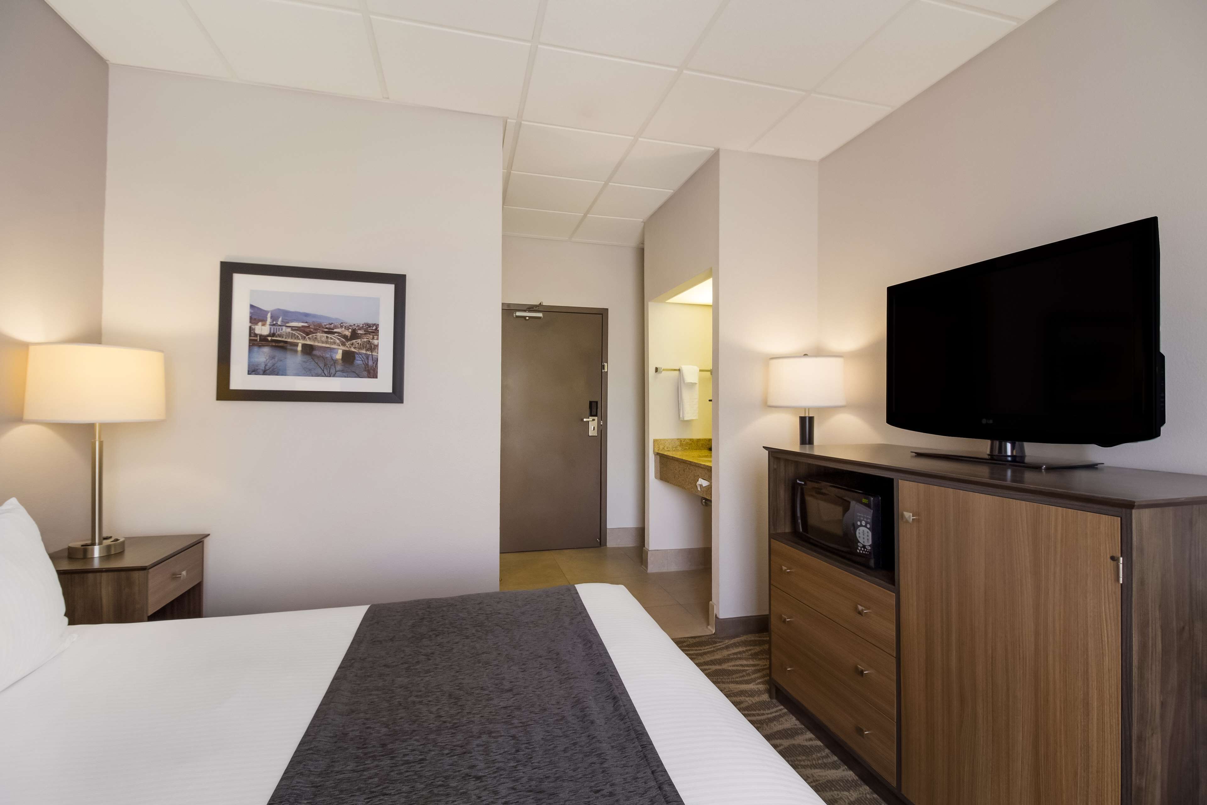 A hotel room with a bed, a television, a desk with a lamp, a framed picture on the wall, and a door leading to a bathroom.