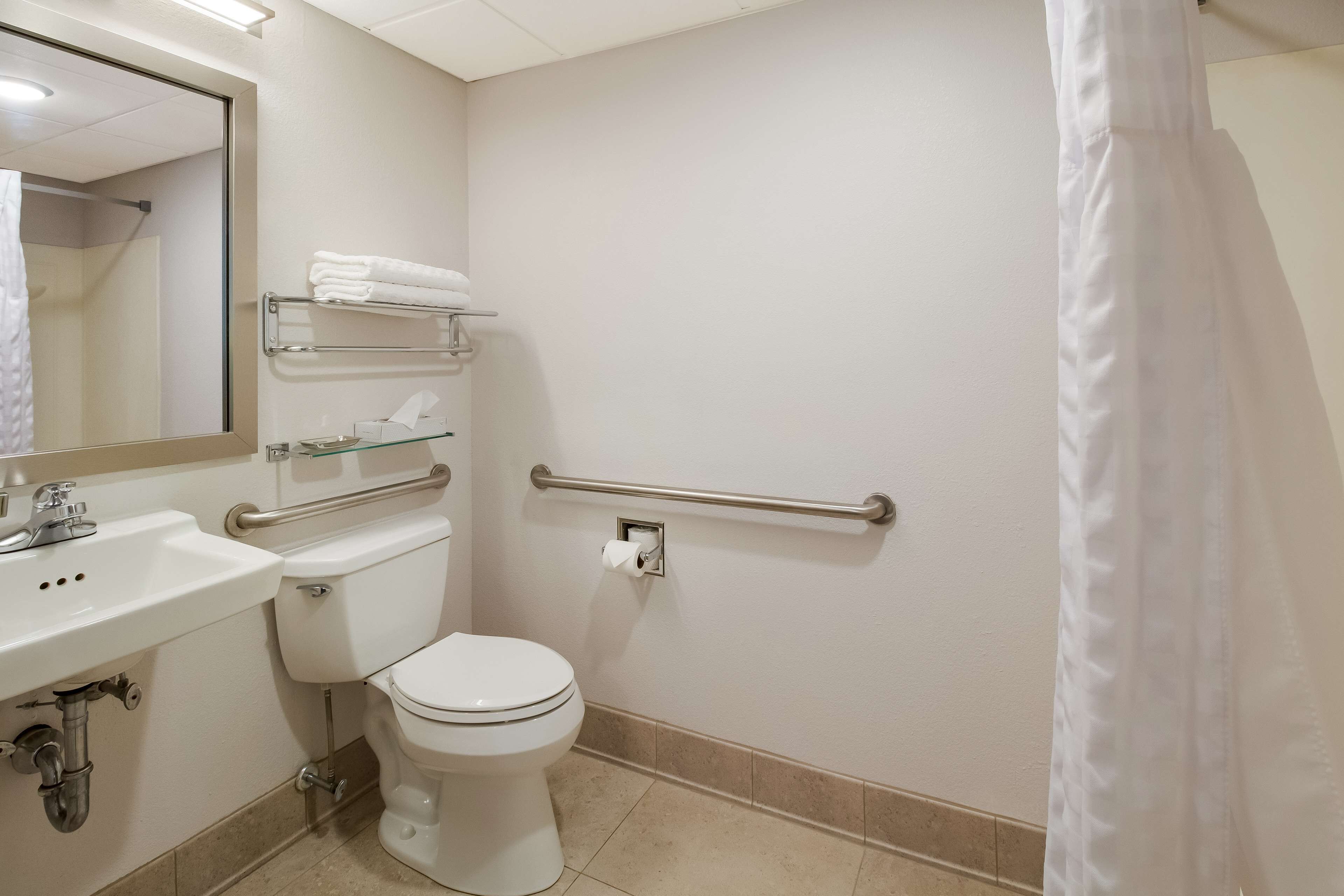 A bathroom with a toilet, sink, and mirror. There is a towel rack above the toilet and a handrail on the wall.