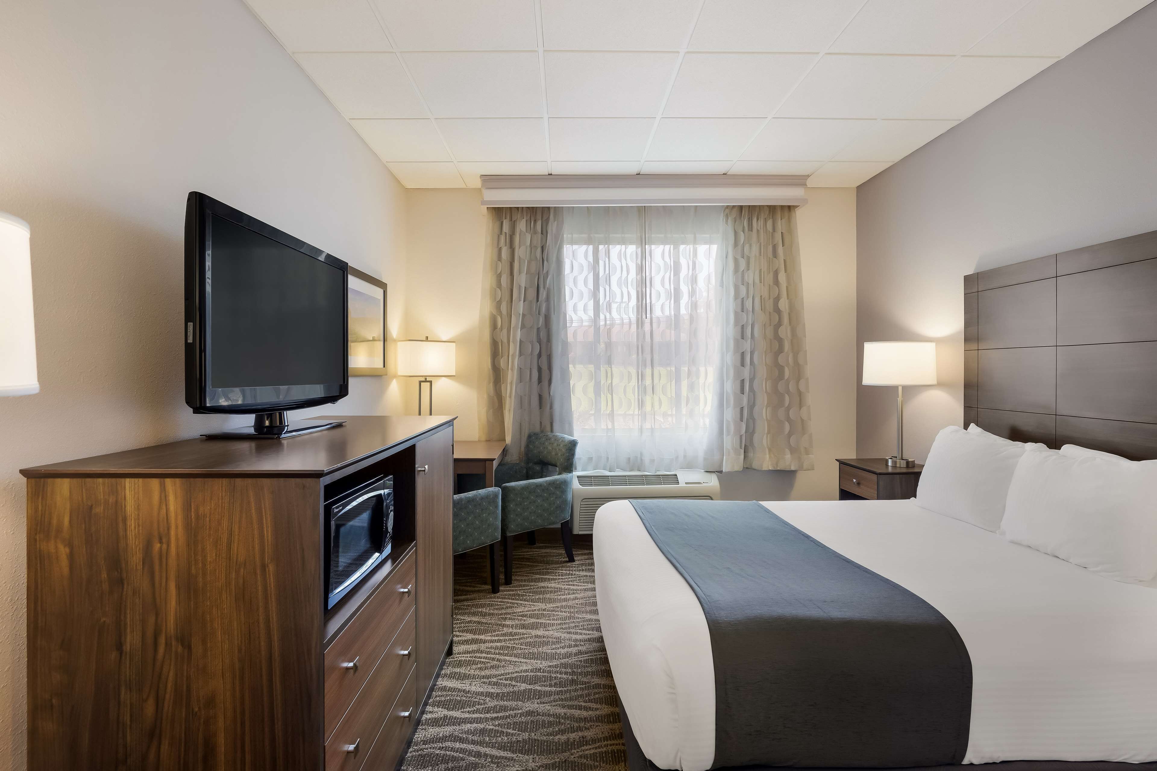 Ein Hotelzimmer mit einem großen Bett, einem Fernseher auf einer Holzkommode, einem Schreibtisch mit Stuhl und einem Fenster mit durchsichtigen Vorhängen.