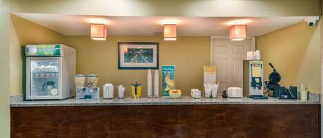 A hotel reception area with a self-service beverage station, including a refrigerator, coffee maker, and various drink dispensers.