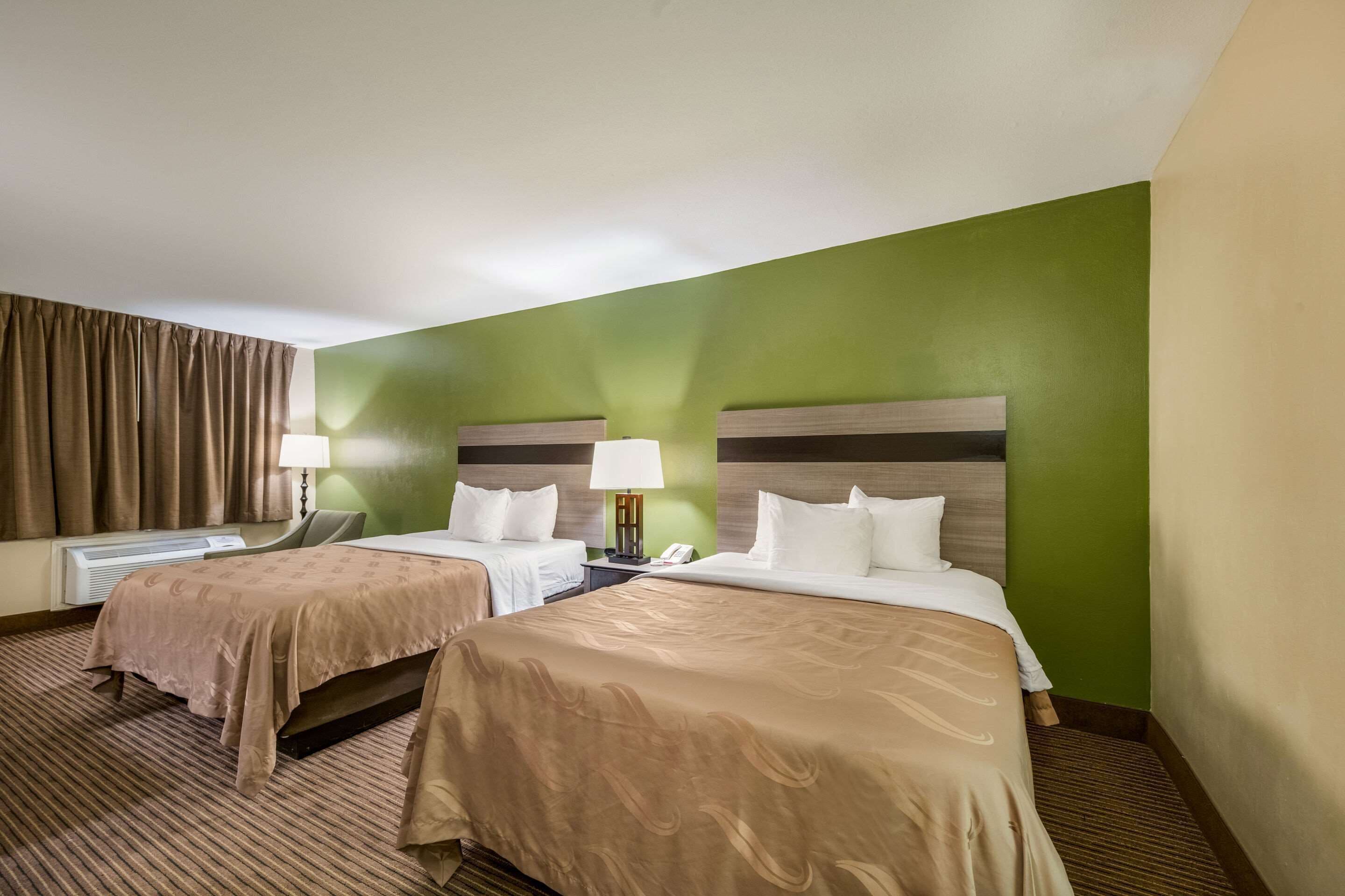 A hotel room with two beds, a green accent wall, a brown carpet, and a window with curtains.