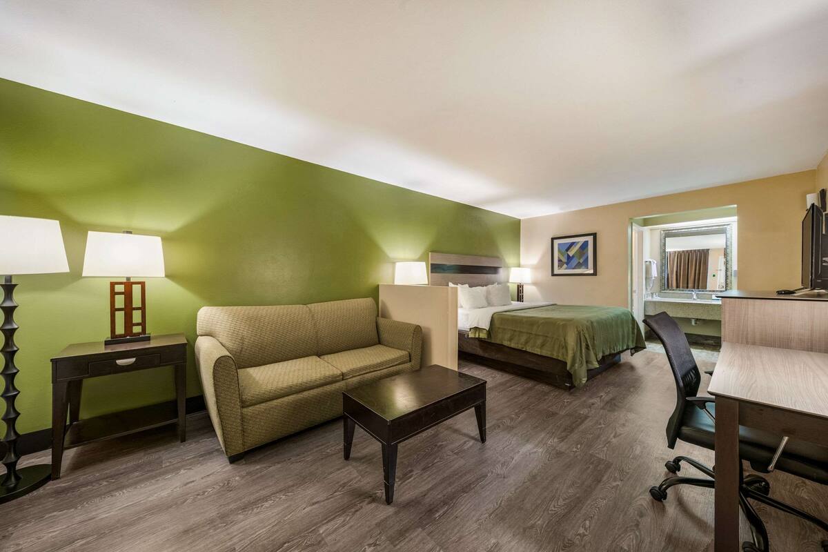 A hotel room with a green wall, a sofa, a bed with a green cover, a wooden coffee table, a desk with a chair, and two bedside lamps.