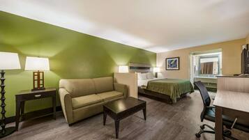 Habitación de hotel con pared verde, un sofá, una cama con cubrecama verde, una mesa de centro de madera, un escritorio con una silla y dos lámparas de noche.