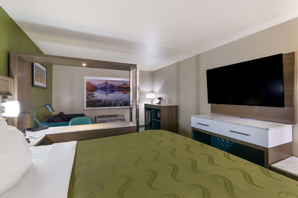 Quarto de hotel com cama, escrivaninha com abajur, televisão e um quadro na parede.