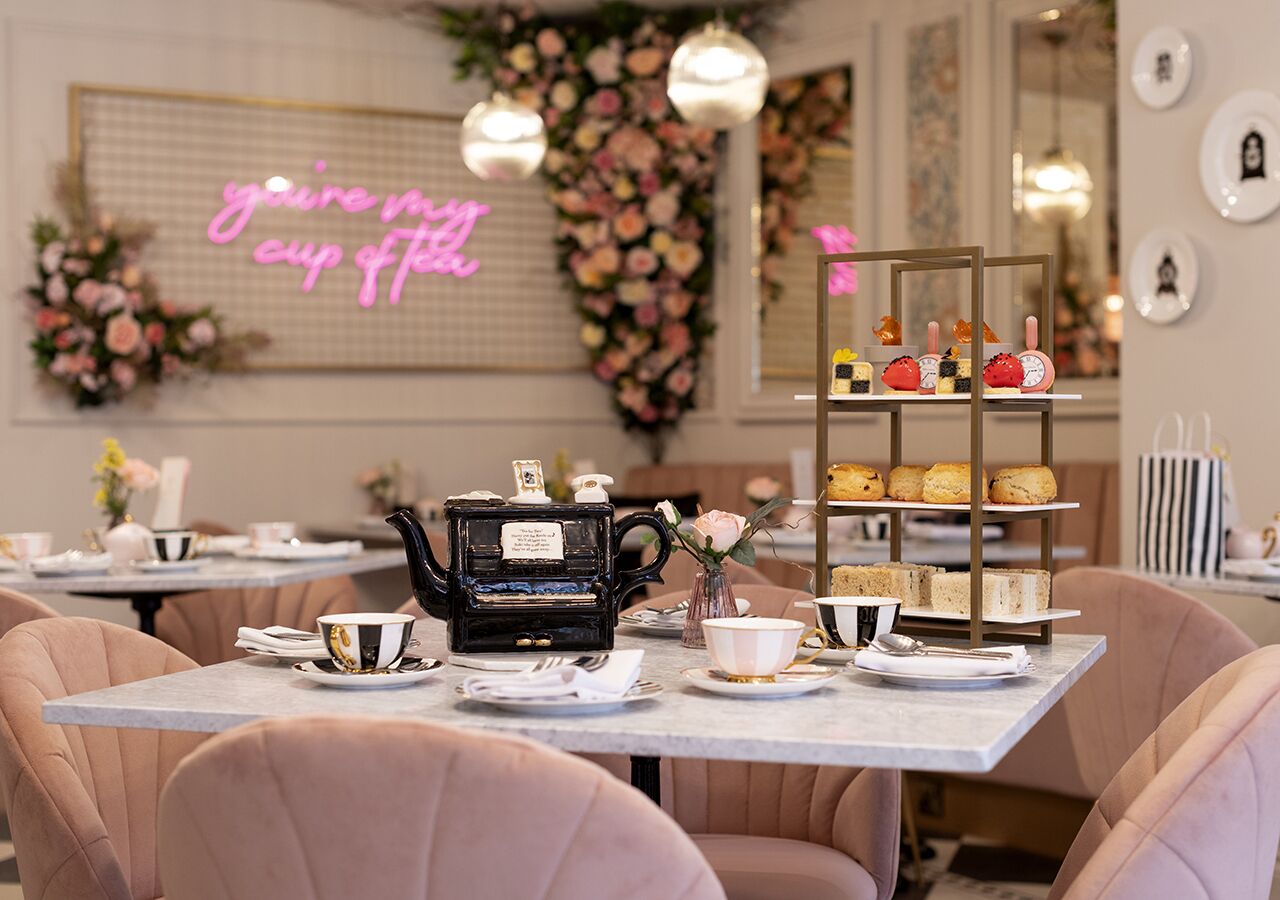 A tea service setup with a teapot, cups, and a tiered display of pastries on a table in a room with floral decorations and a neon sign.