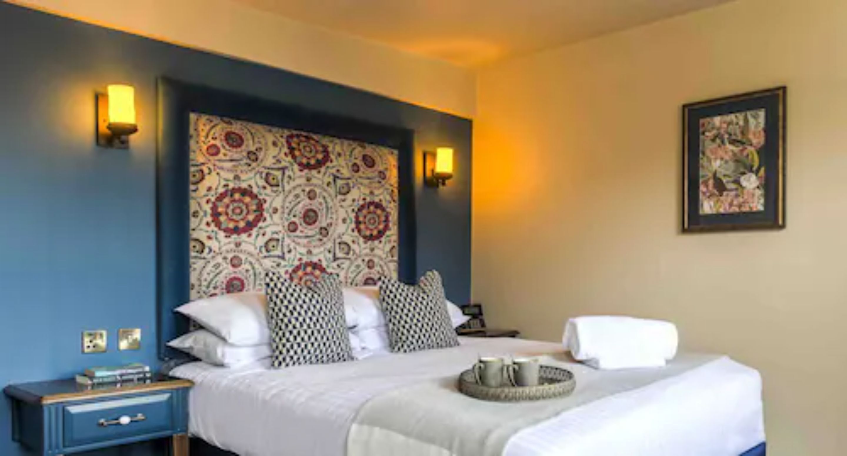 A hotel room with a bed, bedside tables, a headboard with a patterned design, wall-mounted lights, and a framed picture on the wall.