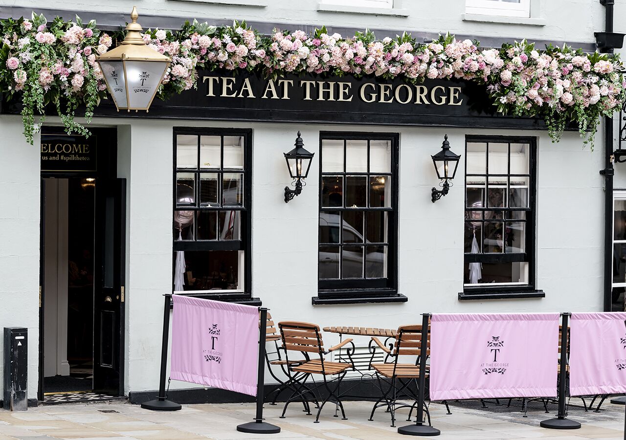 Un edificio blanco con un letrero negro que dice "TEA AT THE GEORGE". Hay dos bancos de madera frente al edificio, y una pancarta rosa con el nombre del edificio está colgada sobre ellos.
