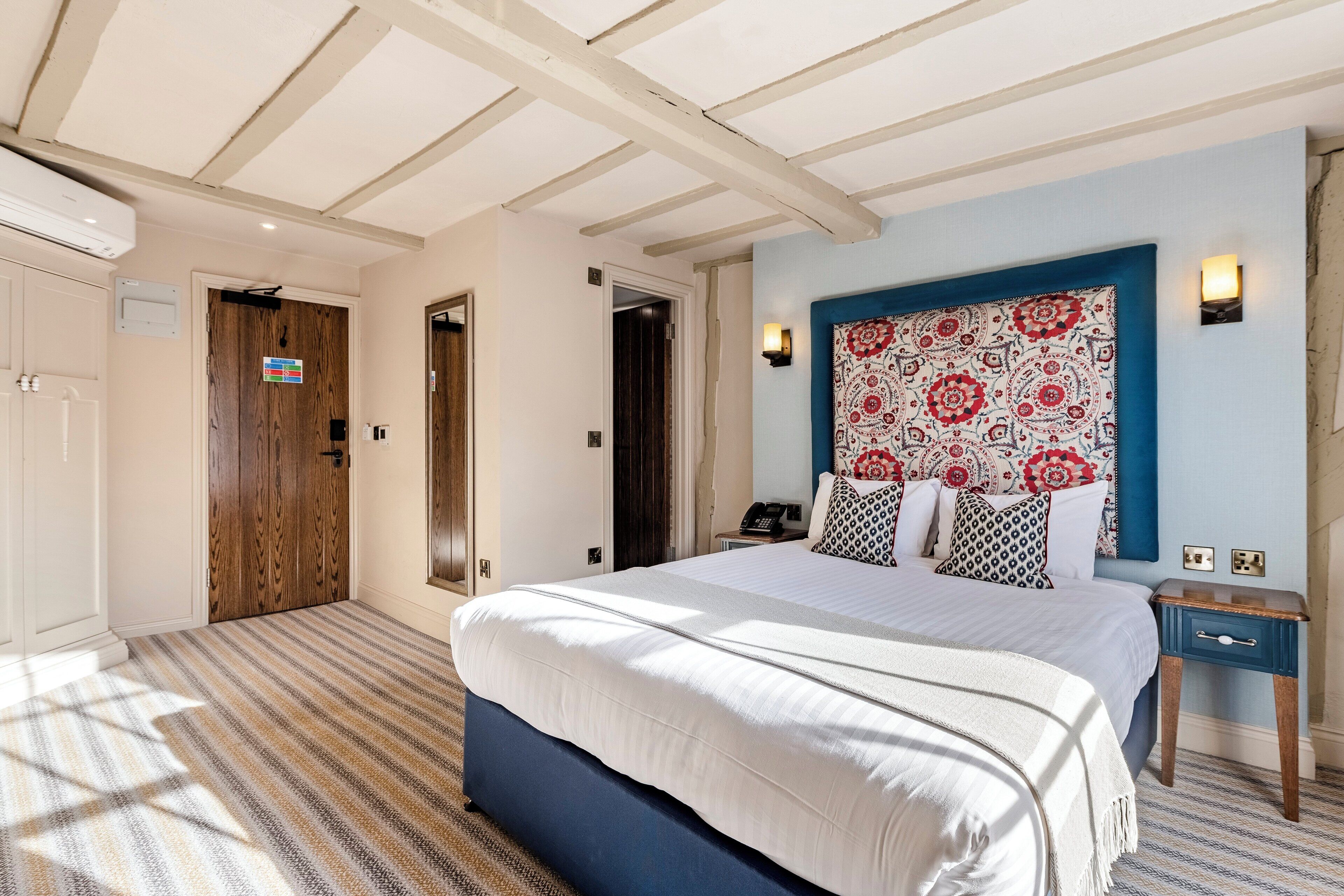 Una habitación de hotel con una cama grande, cabecera con diseño estampado, mesita de noche con lámpara y puerta con un cartel.