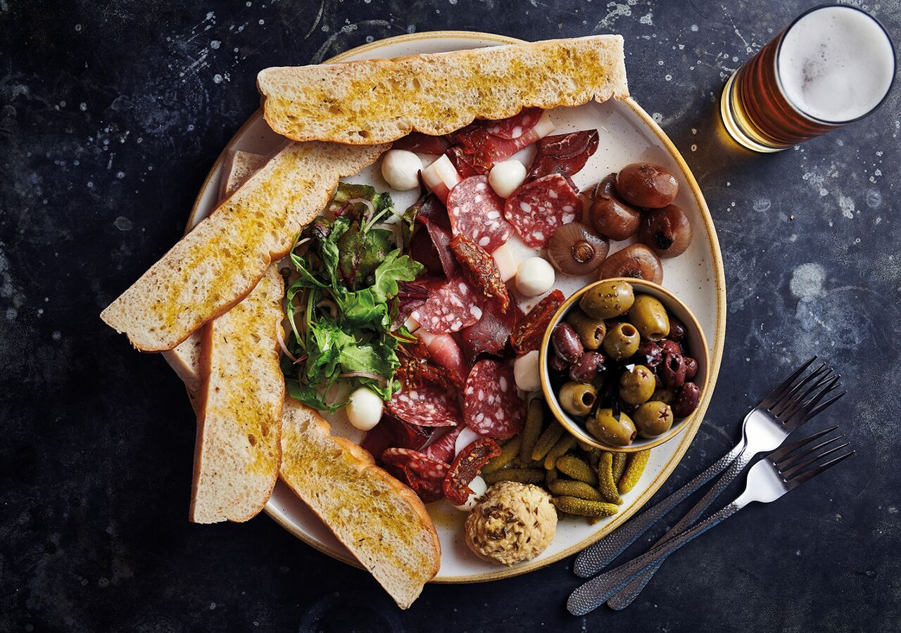 Un plato con diversos embutidos, aceitunas, encurtidos y pan sobre una superficie oscura.