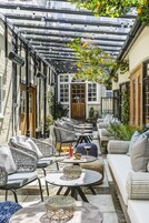 A covered outdoor seating area with wicker furniture and a glass table, surrounded by stone walls and a wooden door.