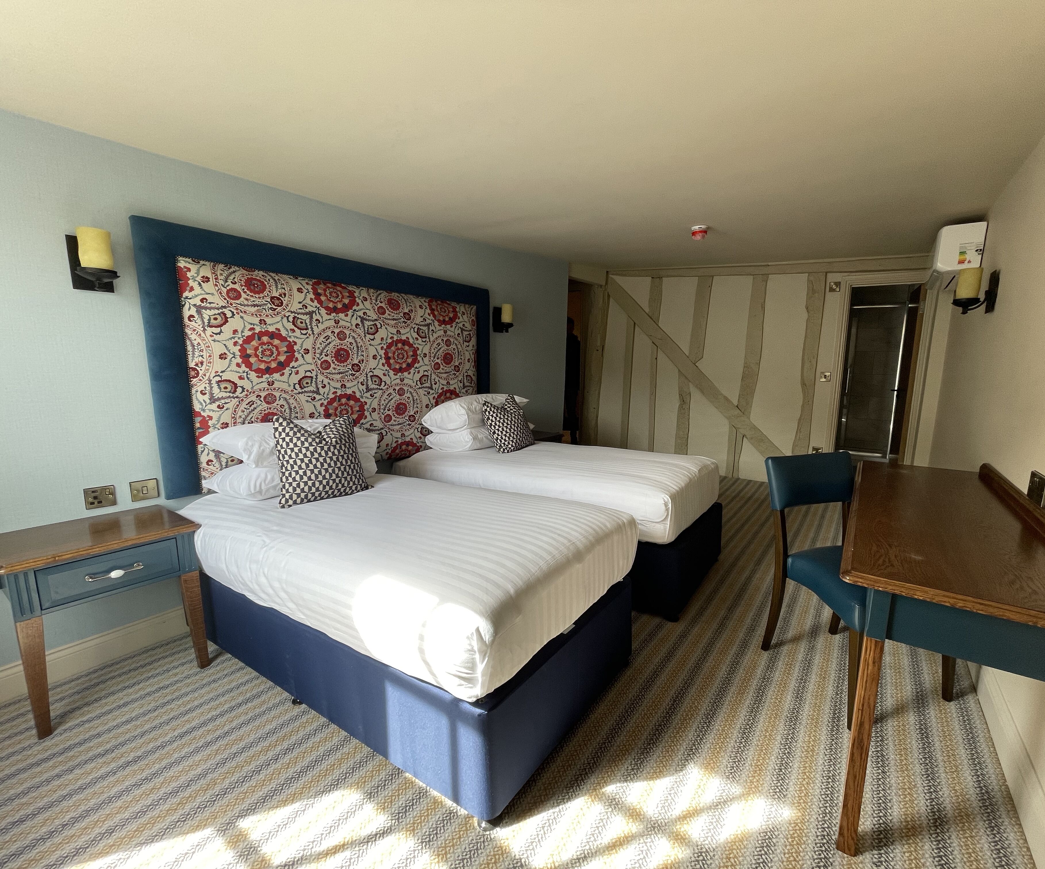 A hotel room with a large bed, a desk, and a chair. There is a patterned wall behind the bed and a window with a view of the outside.