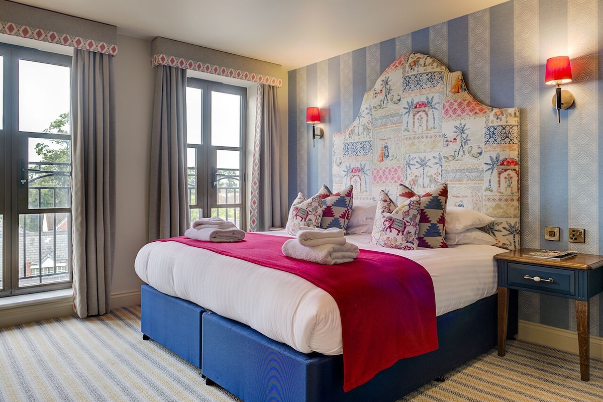 A hotel room with a large bed, a nightstand, a window with curtains, and a patterned headboard.