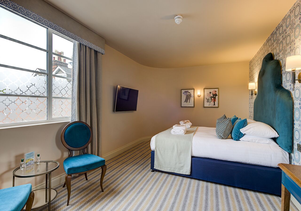 A hotel room with a bed, a chair, a small table, and a window with curtains.
