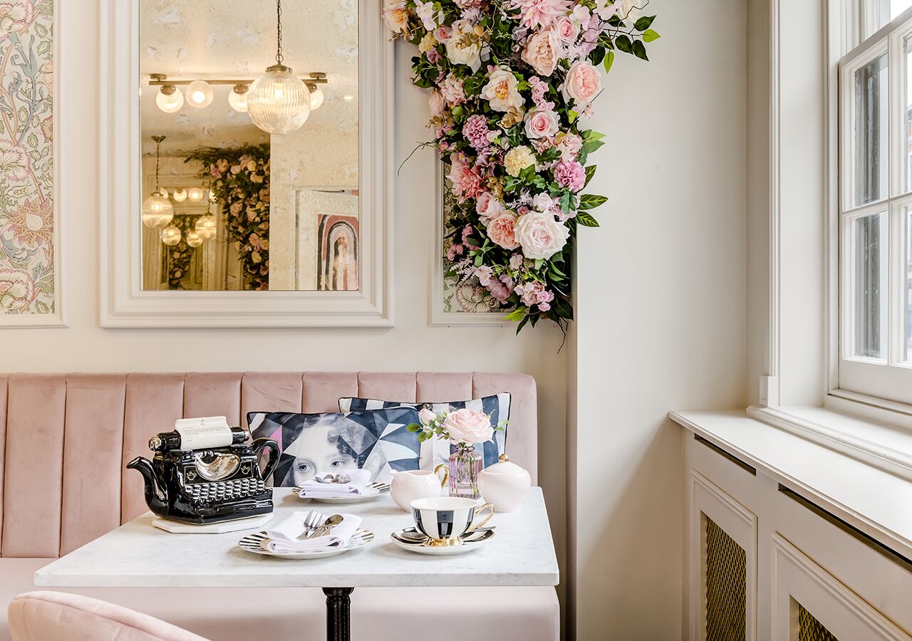 A dining area with a table set for tea, a pink cushioned bench, a mirror, and a floral wall decoration.