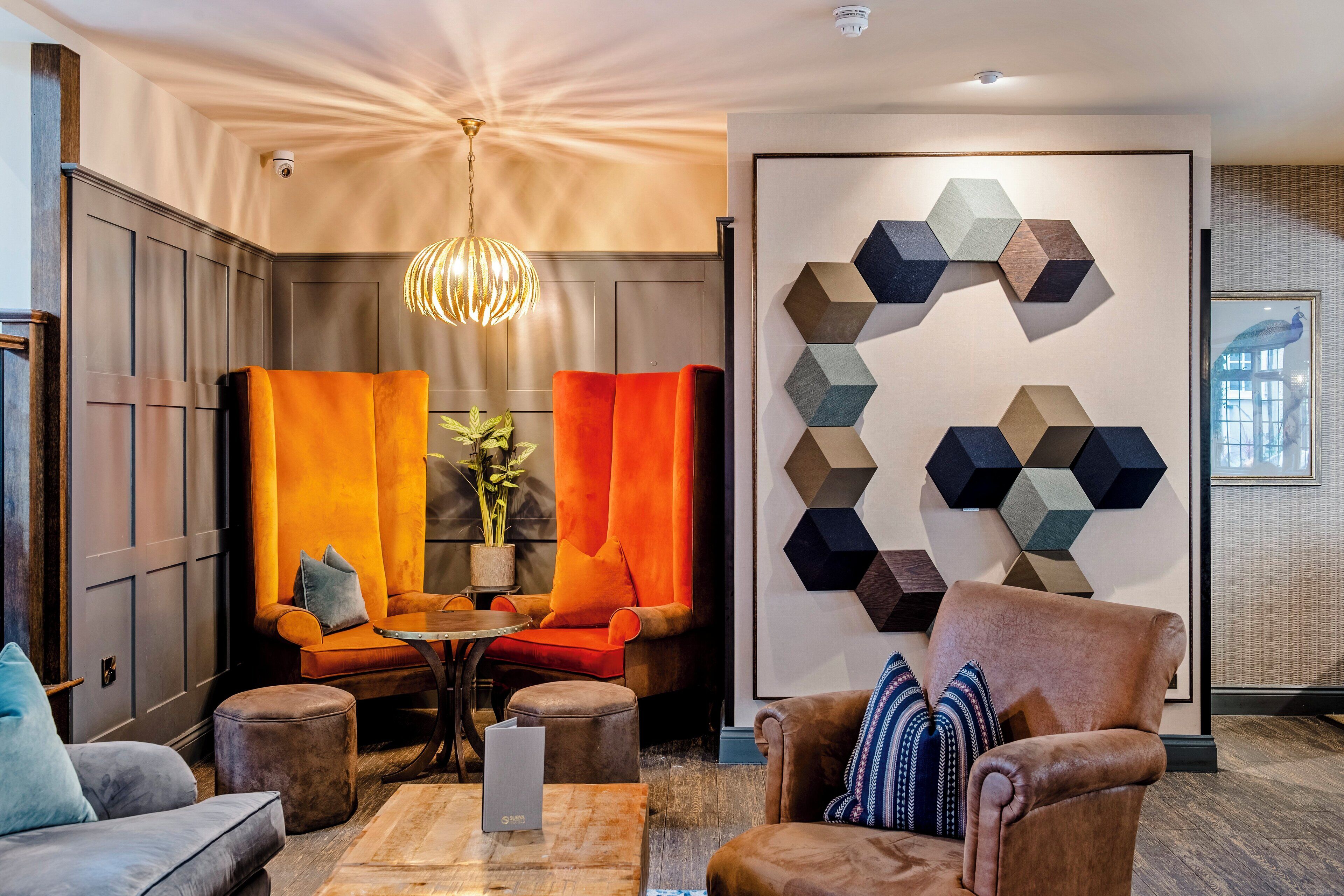 A modern lounge area with a seating arrangement including a sofa, armchairs, and ottomans. There is a coffee table in the center, a potted plant, and a wall-mounted geometric art piece.