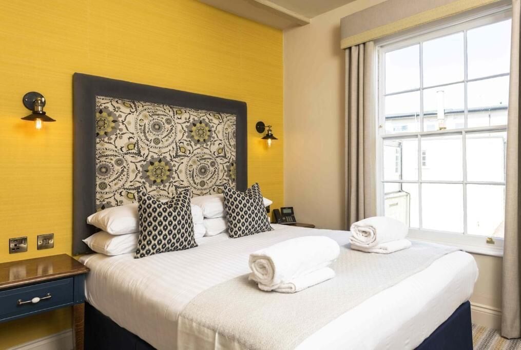 A hotel room with a large bed, patterned headboard, bedside lamps, a window with curtains, and a neatly made bed with towels.