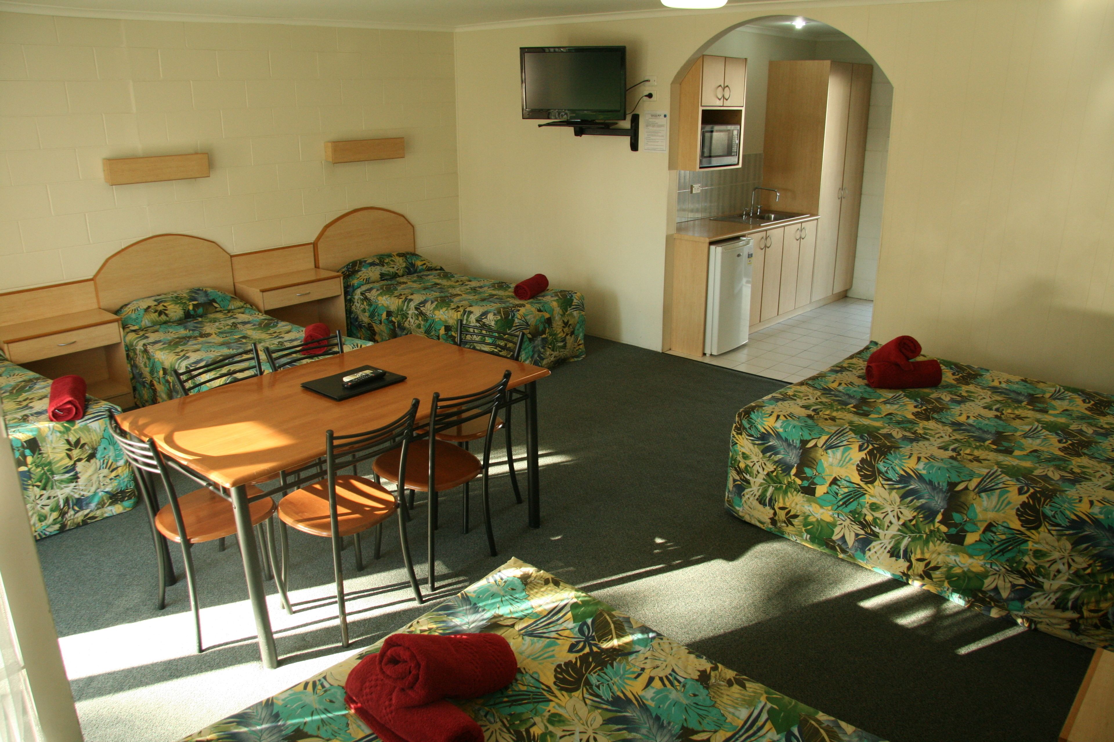 A compact room with a dining table, two single beds, a kitchenette, and a TV mounted on the wall.