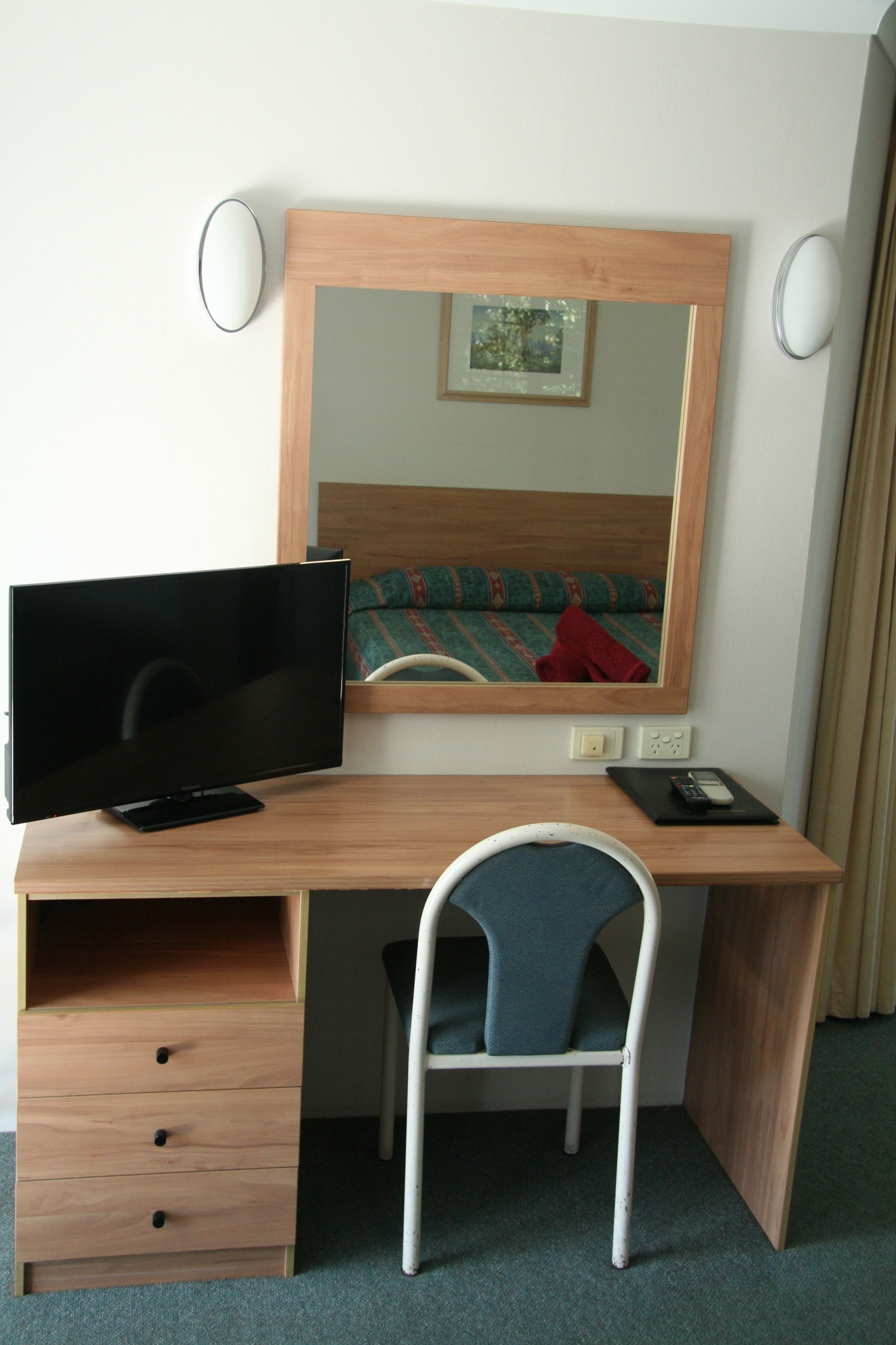 A hotel room with a wooden desk, a television, a chair, a mirror, and a bed with a red blanket.