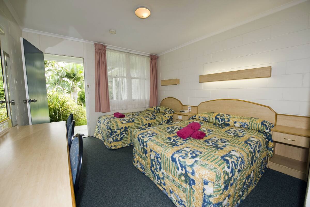 A hotel room with two beds, a wooden desk, a small bedside table, and a window with curtains.