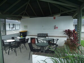A covered outdoor seating area with tables and chairs, a grill, and a trash bin.