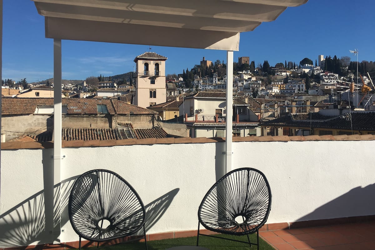 Un balcón con dos sillas, un muro blanco y vistas de un paisaje urbano con edificios y una torre.