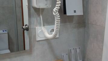 A bathroom with a hairdryer mounted on the wall, a mirror, a white shelf with two glasses, a basket with toiletries, and a whiteboard.