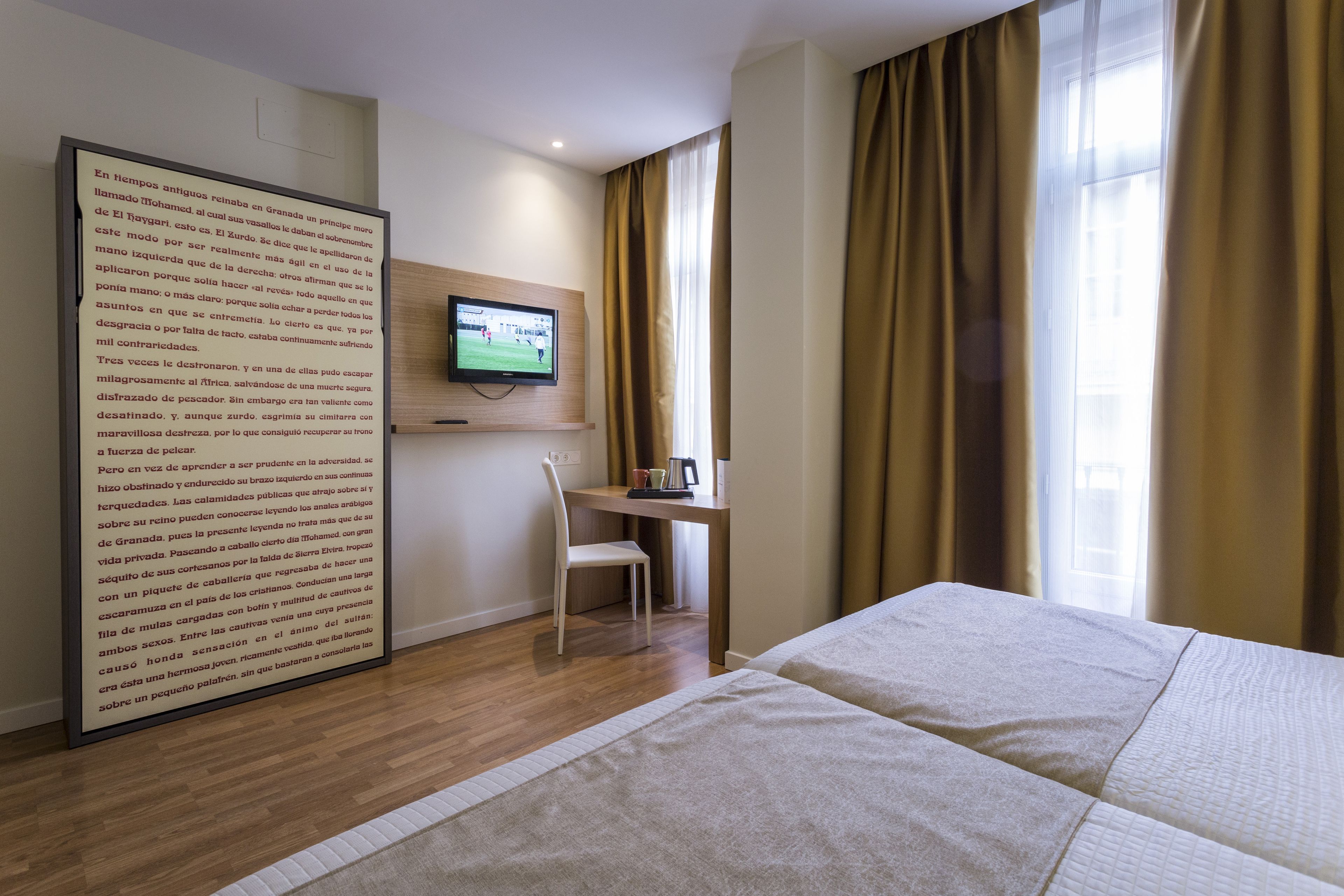 Une chambre d’hôtel comprenant un lit, un bureau avec une chaise, un téléviseur fixé au mur et une grande fenêtre avec des rideaux.