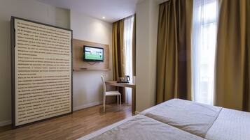 Habitación de hotel con cama, escritorio con silla, televisor en la pared y un ventanal con cortinas.