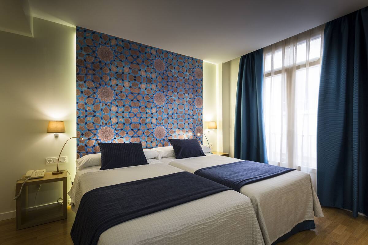 Habitación de hotel con dos camas, un papel tapiz estampado, una mesita de noche con teléfono y una ventana con cortinas.