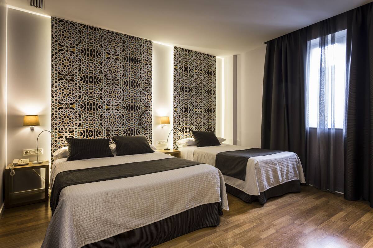 Habitación de hotel con dos camas, un papel tapiz decorativo, una mesita de noche con lámpara y una ventana con cortinas.