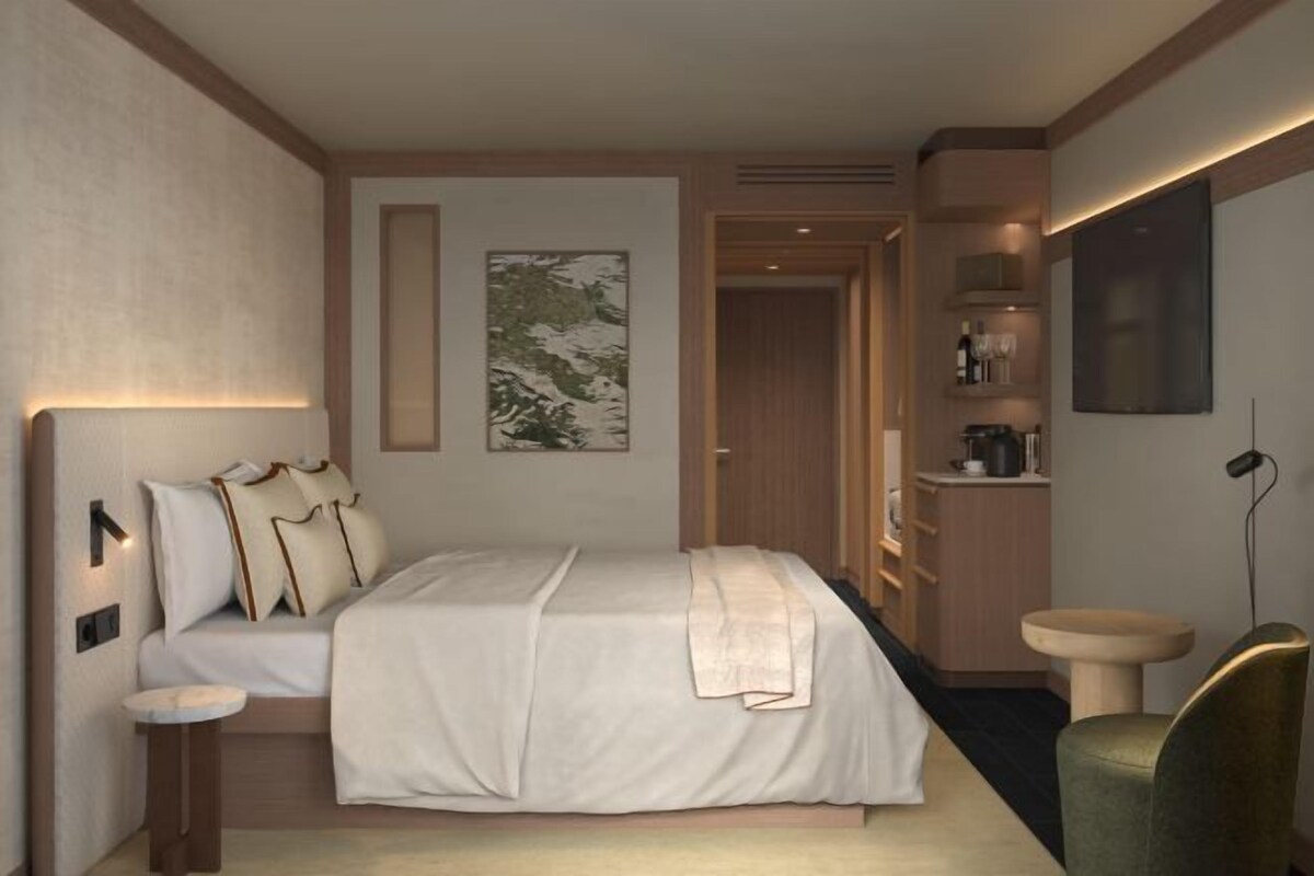 Una habitación de hotel con cama, mesita de noche, lámparas de pared, un cuadro, baño con lavamanos y una silla.