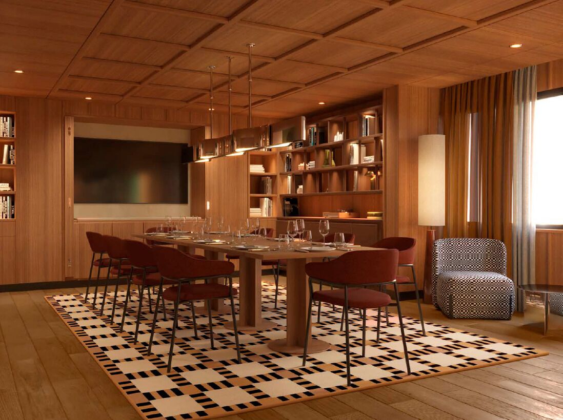 A modern dining area with a large table, red chairs, and a patterned rug. There are bookshelves and a large screen in the background.
