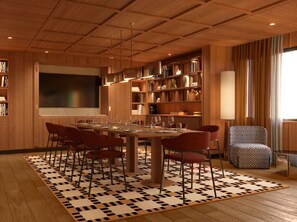 Un espace repas moderne avec une grande table, des chaises rouges et un tapis à motifs. On aperçoit des étagères et un grand écran au fond.