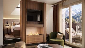 A modern hotel room with a fireplace, a green armchair, a coffee table, and a view of mountains through large windows.