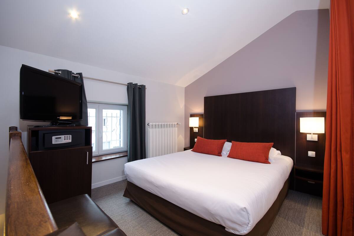 Hotelkamer met een groot bed, een televisie op een houten dressoir, een raam met gordijnen en twee bedlampjes.
