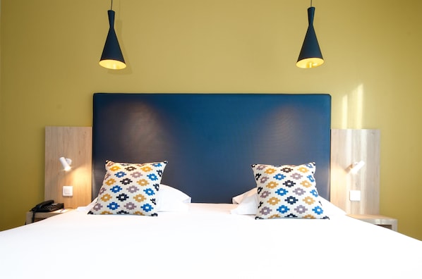 Habitación de hotel con una cama grande, dos almohadas estampadas, un cabecero de madera, lámparas de noche y un teléfono en la mesita.