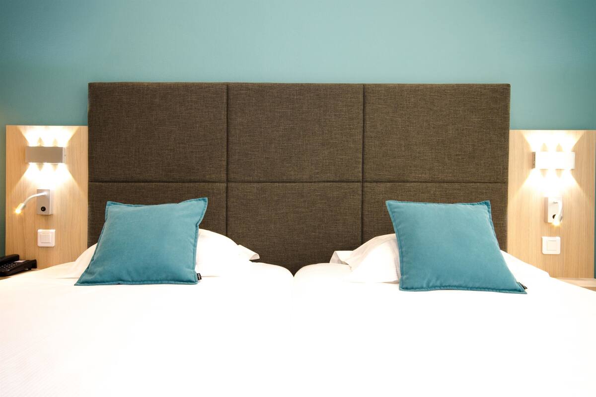 Een hotelkamer met een groot bed, twee blauwe kussens, een bruin hoofdeinde en wandlampen.