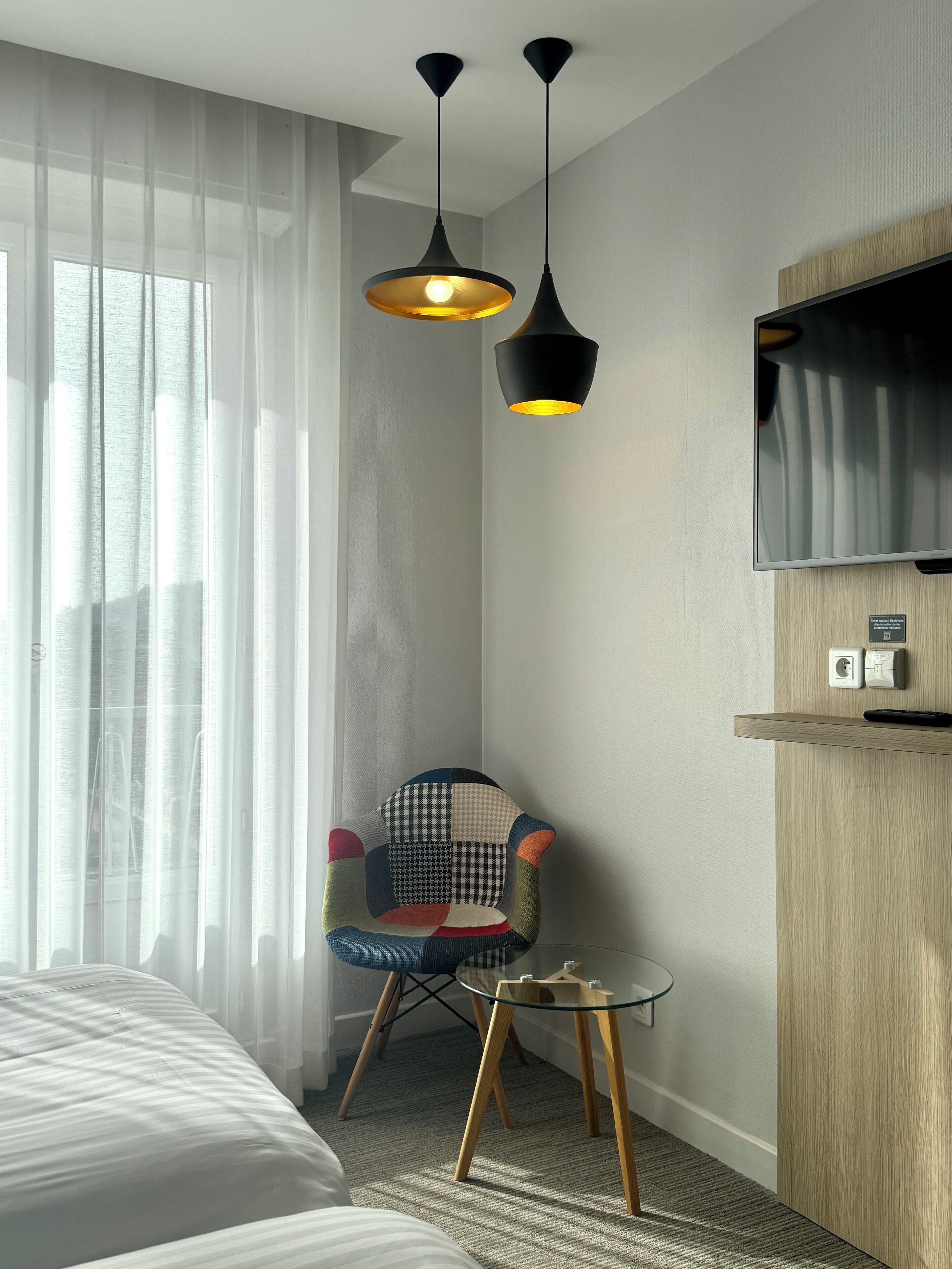 A hotel room with a bed, a chair, a small table, a TV mounted on the wall, and sheer curtains.