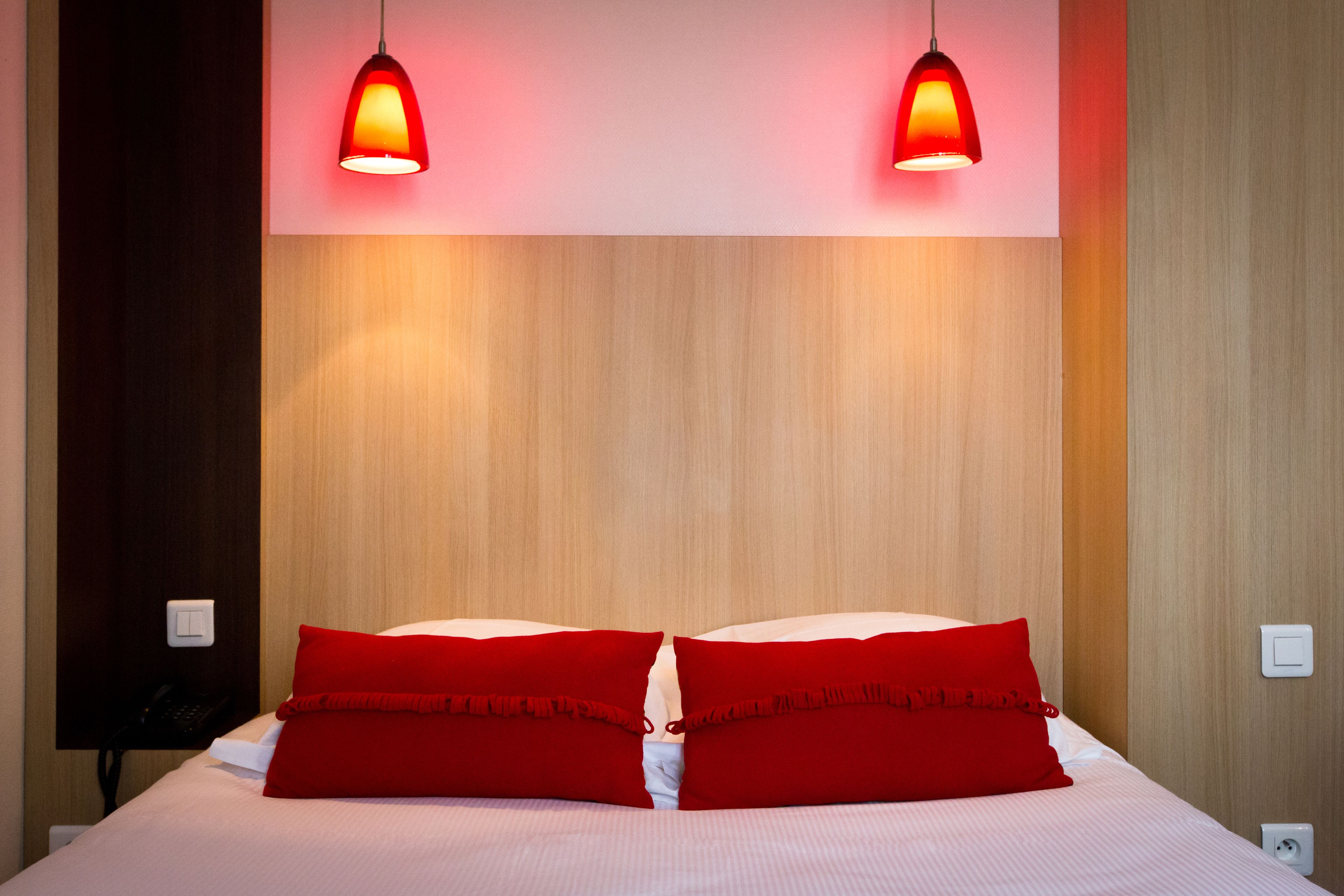 Habitación de hotel con una cama, dos almohadas rojas, un cabecero de madera y dos lámparas colgantes.