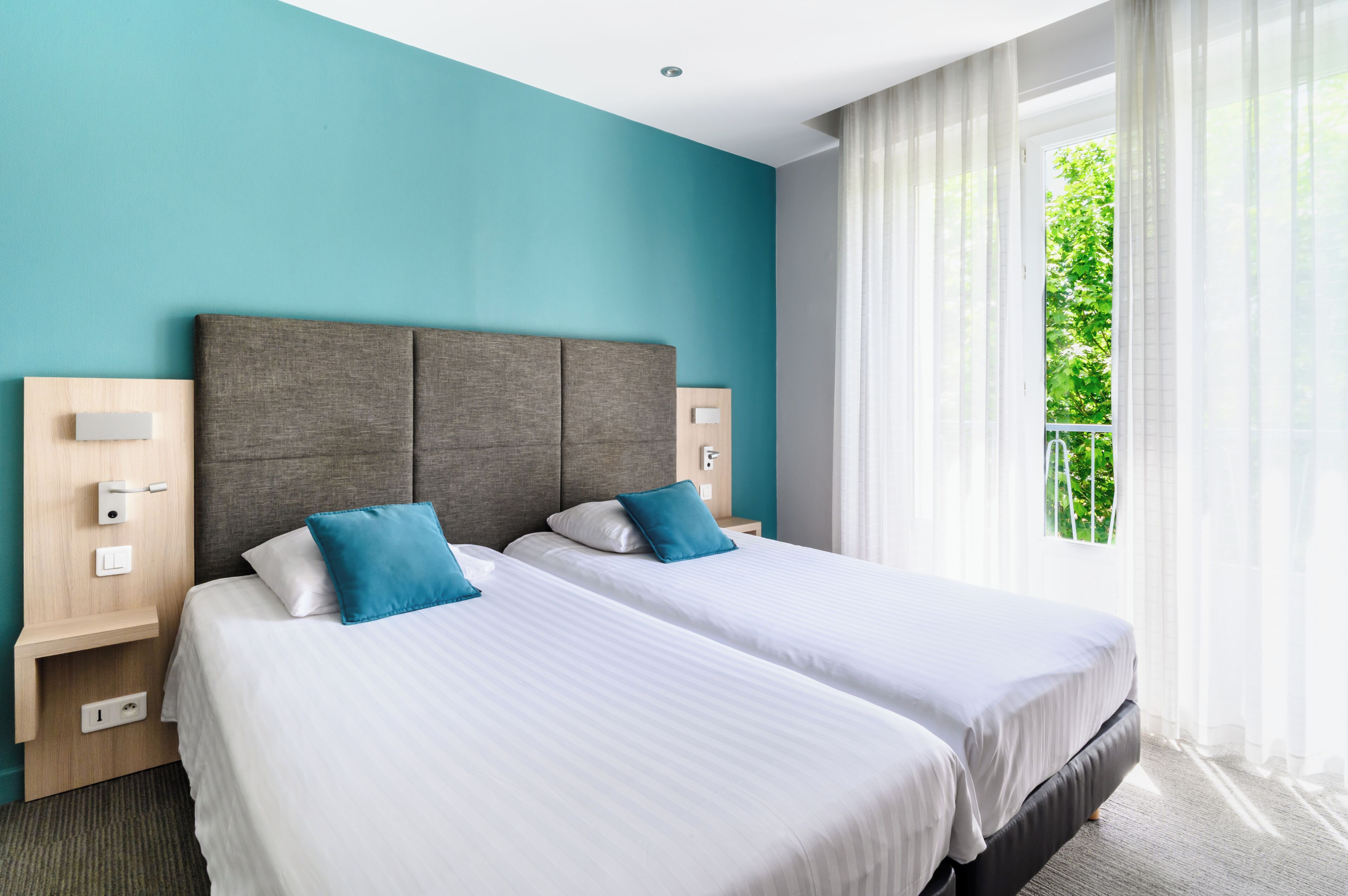 A hotel room with two beds, a teal wall, and a window with a view of greenery.