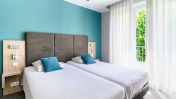 A hotel room with two beds, a teal wall, and a window with a view of greenery.