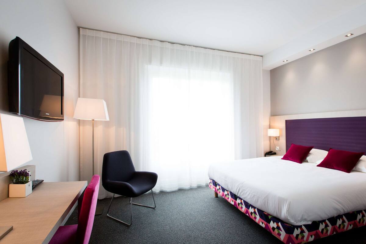 Hotelkamer met een bed, bureau, stoel, televisie en raam met gordijnen.