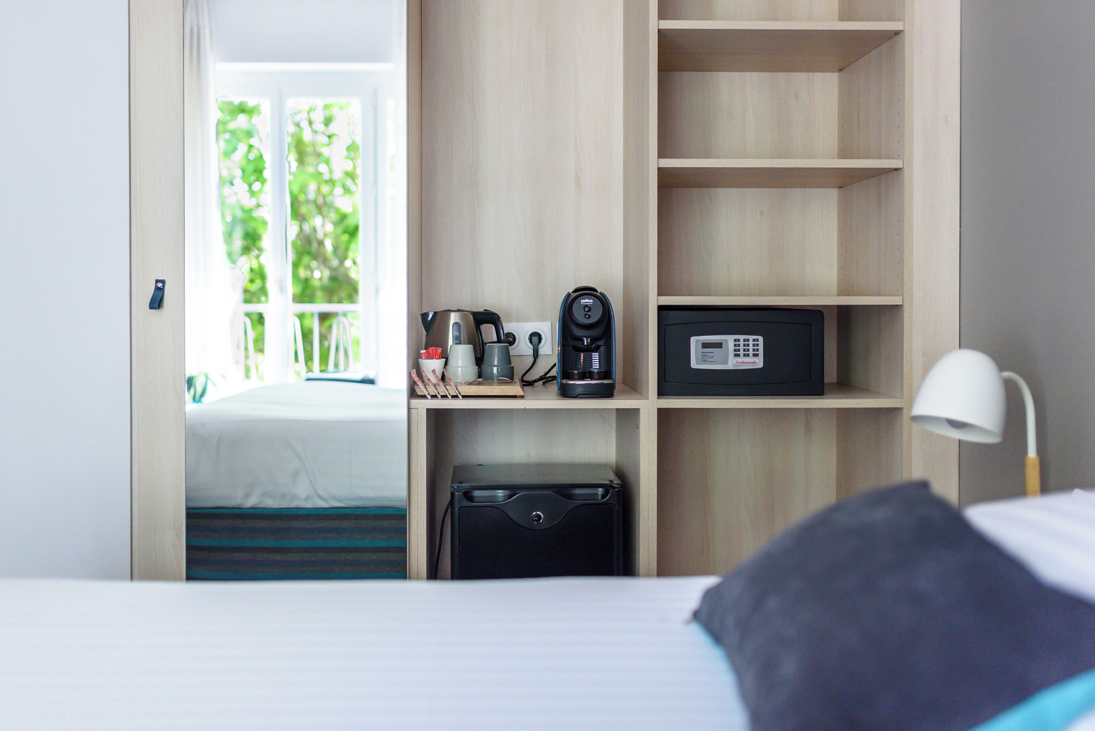 A hotel room with a bed, a desk with a kettle, a radio, and a suitcase.