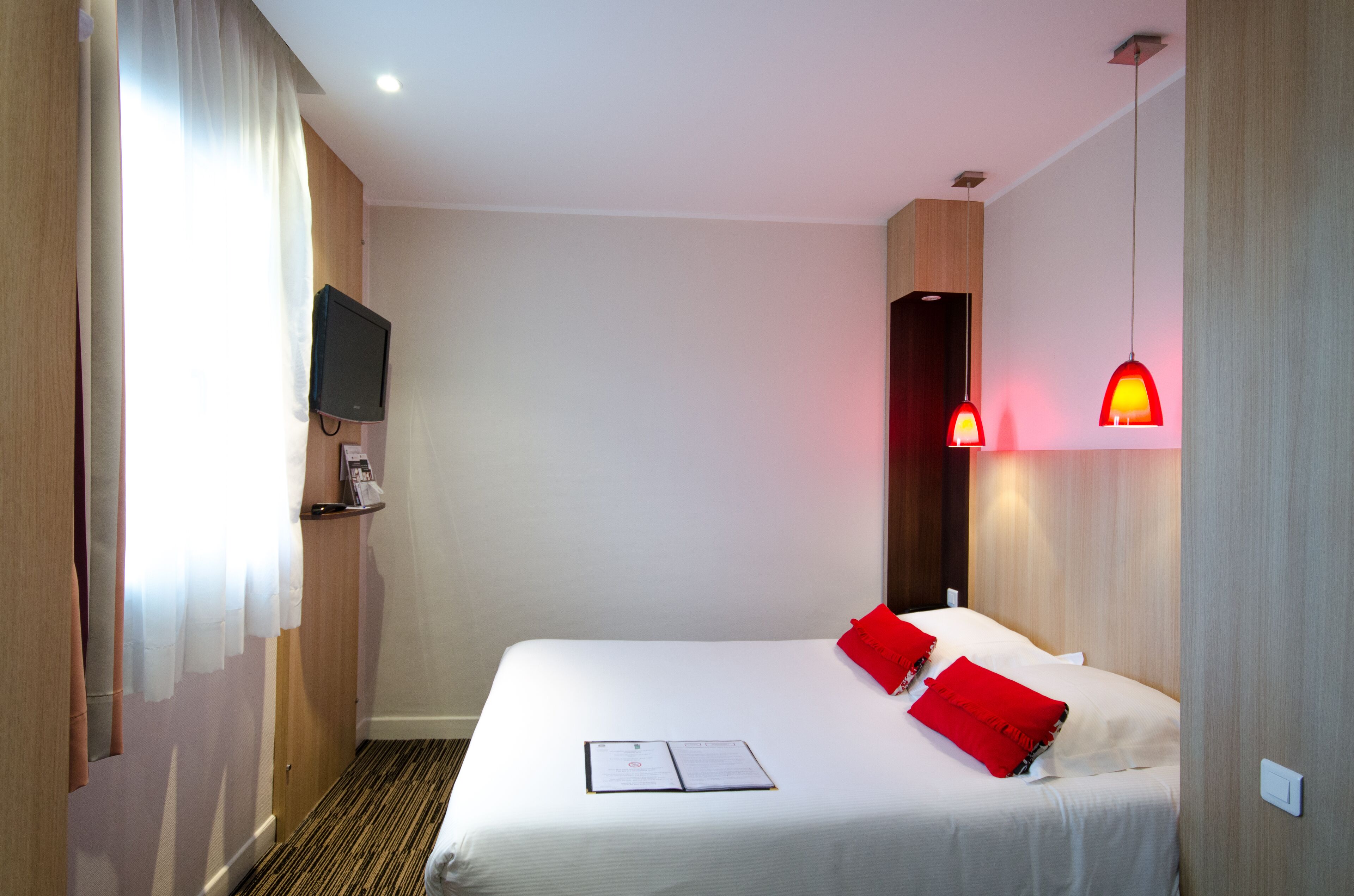 Ett hotellrum med en säng, röda kuddar, en platt-TV och en röd hänglampa.